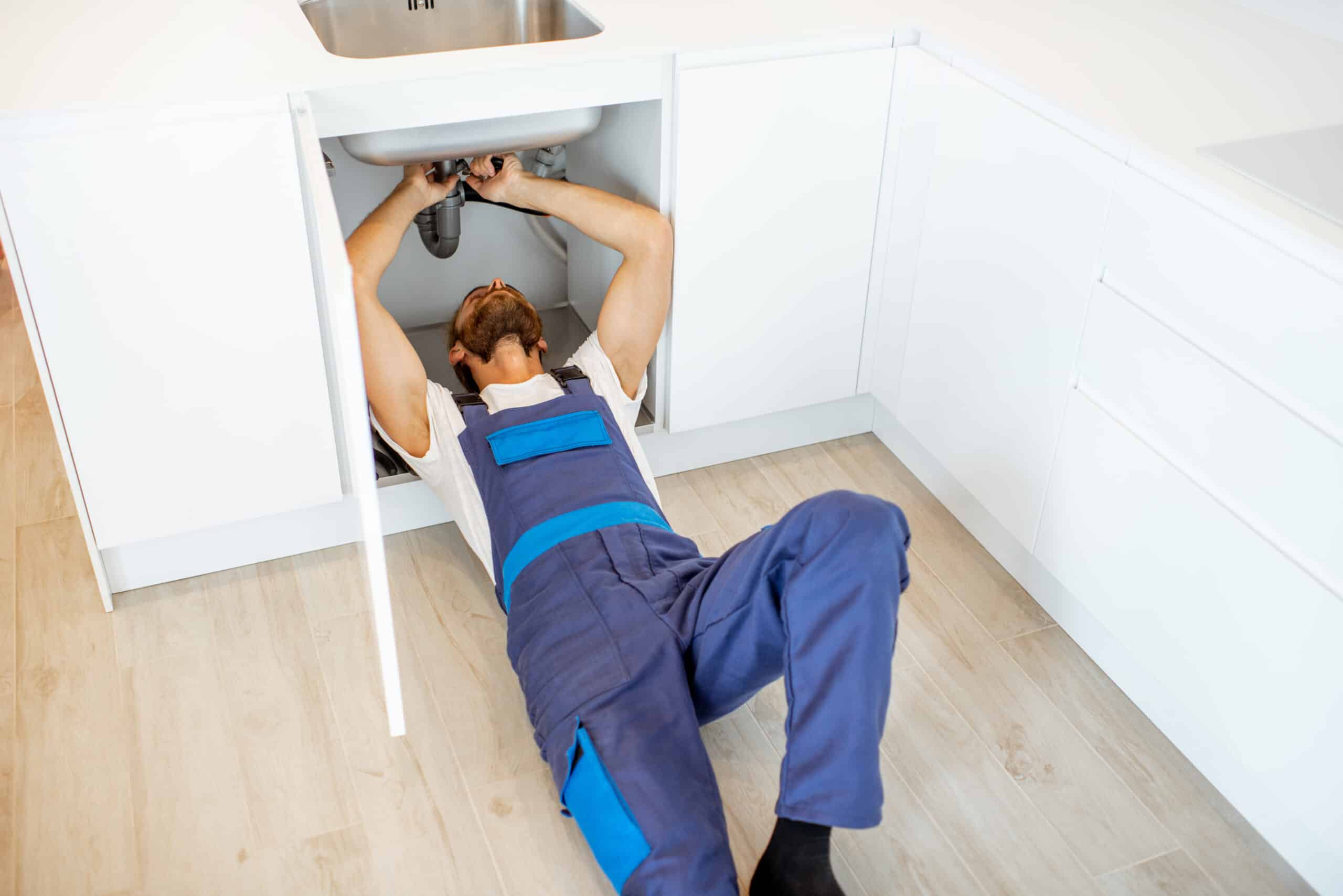 Plumber in overalls repairing or installing sewerage under the kitchen sink at home